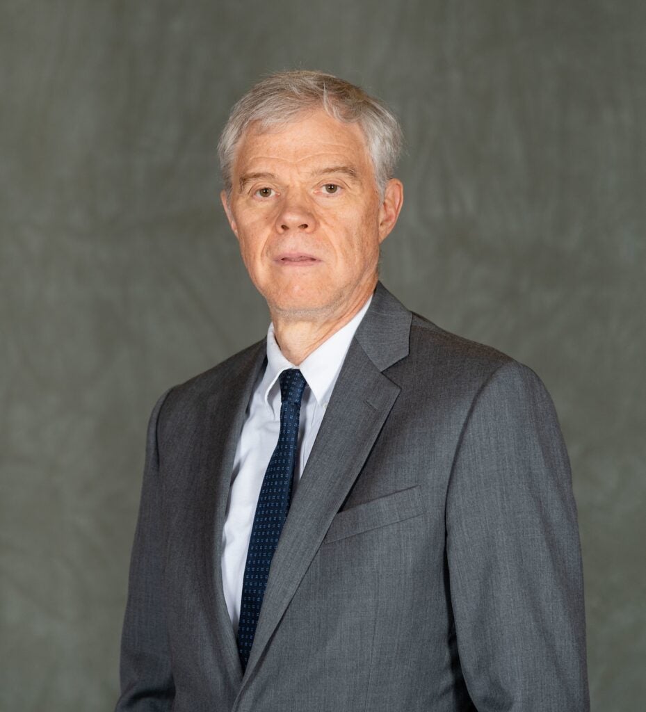 Brian Madigan in a grey suit jacket and navy blue tie against a grey background