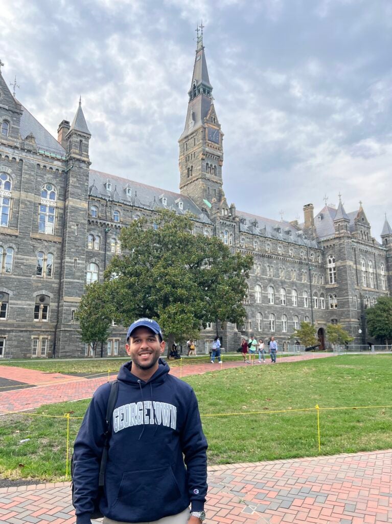 Carlos Segura Santana wearing a Georgetown sweatshirt and baseball cap, standing outside in front of Healy Hall.