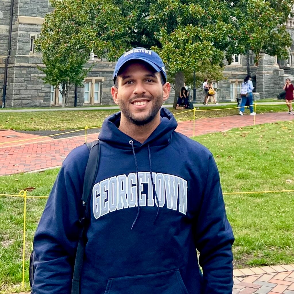 Carlos Segura Santana wearing a Georgetown sweatshirt and baseball cap, standing outside in front of Healy Hall.